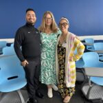 UCLA MHA Program Director Dr. Laura Erskine (center) stands smiling between MHA graduates and guest judges Bryan Ceron (left) and Monique Trinh (right) in a classroom. Bryan is wearing a black long-sleeve shirt, Dr. Erskine is in a green and white floral dress, and Monique is wearing a sunflower-patterned dress with a yellow-and-white striped cardigan. The background features several rows of bright blue modern classroom chairs and a vibrant blue accent wall.