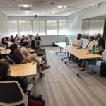 A career panel discussion held in a modern, brightly lit UCLA classroom during the Fall 2025 MHA Immersion. Three panelists—Curana Health COO Craig DePriester, UCLA MPH Graduate and Kaiser-Permanente Regional Workshop Consultant Nikhil Kulkarni, and MHA Graduate and UCLA Bedari Kindness Institute Fellow Monique Trinh—are seated at a long wooden table on the right side of the room, facing a group of approximately 15 students seated at desks. The panelists and students are dressed in professional attire, and a large, dark digital screen is mounted on the wall behind the speakers.