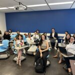 A group of approximately 15 UCLA MHA students seated in a modern classroom during the Fall 2025 Immersion. The students are dressed in professional attire, seated in light-blue swivel chairs with attached desks, and smiling as they prepare for their case competition presentations. The room features a vibrant blue accent wall and a ceiling-mounted projector.