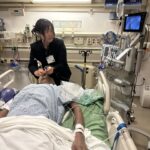 A UCLA MHA student in a black blazer participating in a hands-on clinical simulation at the Cedars-Sinai Women’s Guild Simulation Center. She is leaning over a medical mannequin in a hospital bed, carefully practicing a procedure with a clinical instrument while looking at a digital monitor displaying a live endoscopic feed. The room is a realistic medical environment equipped with vital sign monitors and wall-mounted hospital equipment.