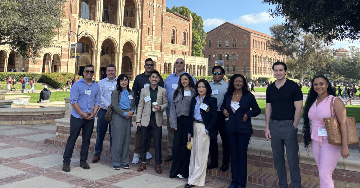 A diverse group of twelve professionally dressed men and women MHA students, many wearing UCLA MHA name tags, pose for a group photo on the UCLA campus. They are standing on a brick landing with the iconic twin-towered, Romanesque-style Royce Hall and a clear blue sky in the background.