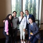 A group of five professionally dressed UCLA MHA students posing for a photo during the MHA Fall 2025 Immersion Farewell Reception at the UCLA Luskin Conference Center. The group is positioned in front of a large window overlooking tall green trees. MHA Student Francis Yau wears a plaid blazer and tie in the foreground holding a glass, while the others stand behind in business casual attire.