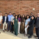 A diverse group of eleven MHA students stand in a row on a sunny patio of UCLA Luskin Conference Center in front of a red brick wall during the Fall 2025 Immersion Farewell Reception at UCLA. The group is dressed in professional business attire—including blazers, slacks, and dresses—and several individuals are wearing official UCLA MHA name tags.