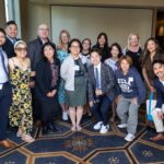 A diverse group of approximately 15 MHA students and faculty smiling and posing together during the Fall 2025 Immersion Farewell Reception at the UCLA Luskin Conference Center. The group is dressed in professional and business casual attire, with several individuals wearing UCLA name tags, standing in a brightly lit room with large windows and a blue and gold geometric patterned carpet.