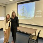 UCLA MHA Student Affairs Officer Jenna Mendoza (left) and Krisianna Bock, Principal with HKS, Inc. (right), posing together in a classroom during the Fall 2025 MHA Immersion. Behind them, a projector screen displays a presentation slide titled “CAREER PERSPECTIVES” with a modern architectural rendering of a healthcare facility. Jenna is dressed in a white knit cardigan and tan trousers, and Krisianna is wearing a black blazer and dark pants.