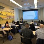 MHA Program Director Dr. Laura Erskine facilitating student introductions during the Fall 2025 MHA Immersion at UCLA. She stands at the front of a classroom next to a projector screen displaying a "Student Bios" slide. The room features a large, vibrant mural of African savannah wildlife on the left wall, and students are seated at light-wood desks with blue UCLA bags.