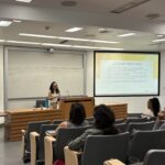 MHA graduate Monique Trinh presenting to a classroom of students during the Fall 2025 MHA Immersion at UCLA. She is standing at a wooden podium in a modern lecture hall, smiling and engaging with the audience. Behind her, a large projector screen displays a slide titled "LEARNING OBJECTIVES" which outlines key takeaways for the session.