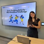 ExecHPM Program Coordinator Sarah Tran stands smiling in front of a large digital display during the MHA Fall 2025 Immersion. The screen features the UCLA Master of Healthcare Administration logo and is titled "Team Building Activity: Marshmallow Challenge" with colorful illustrations of figures in motion. A laptop is visible on a light-wood table in the foreground.