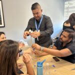 UCLA MHA student Bosco Do carefully places a white marshmallow atop a delicate tower constructed of uncooked spaghetti and blue painter's tape. Two other students sit at a wooden table, focusing intently and using their hands to stabilize the base of the tower. Bosco is wearing a dark grey button-down shirt and a blue UCLA Bruins lanyard during this "Marshmallow Spaghetti Tower Challenge" at the Fall 2025 Immersion.