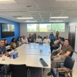UCLA MHA Program Director Dr. Laura Erskine and MHA Student Affairs Officer Jenna Mendoza stand with a group of approximately 15 UCLA MHA students and staff seated around a long, white rectangular conference table during a site visit to Venice Family Clinic. The room features a vibrant blue accent wall with a large mounted monitor and bright windows in the background showing outdoor greenery. The students are dressed in business casual attire and engaged in a group discussion.