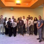 UCLA MHA students pose for a photo with Venice Family Clinic CEO Dr. Mitesh Popat during the Fall 2025 Immersion. The diverse group of about 14 individuals is standing in a modern clinic hallway with pale yellow walls and a bright orange ceiling cove. Everyone is dressed in professional business casual attire, and several students are wearing visible UCLA MHA name tags.
