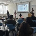 Three UCLA MHA students presenting their project for the mini case competition during the Fall 2025 Immersion. One student in a floral dress stands at the center speaking, while her teammates stand beside her at a podium. Behind them, a projector screen displays a slide titled “INITIATIVE #2: BROADEN & DIVERSIFY COMMUNITY WELLNESS PROGRAMS.” In the foreground, fellow students are seated in blue modern desks, listening to the presentation in a bright classroom.