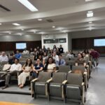 UCLA MHA Graduate Monique Trinh poses with a group of approximately 25 UCLA MHA students in a modern, tiered UCLA lecture hall during the Fall 2025 MHA Immersion. Monique is seated in the front row, second from the left, wearing a checkered sleeveless dress and glasses. The diverse group of students is dressed in professional business casual attire and smiling for the commemorative photo. In the background, two wall-mounted digital screens display presentation slides.