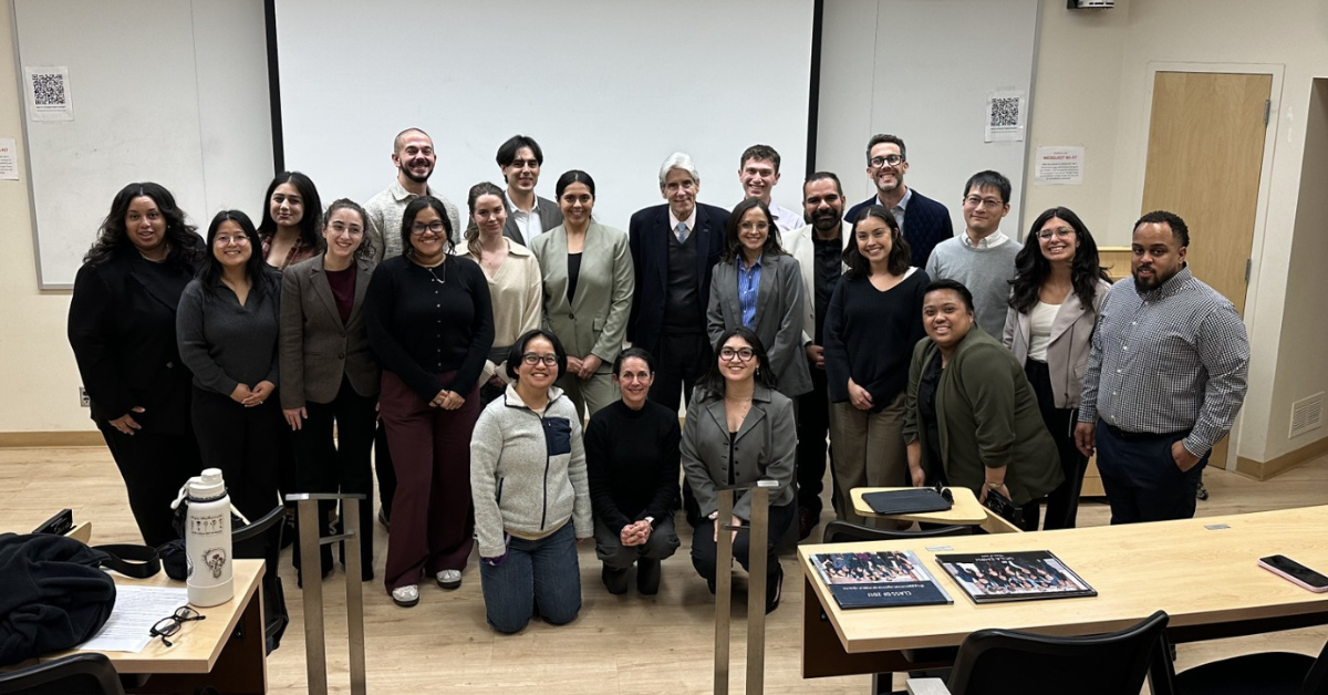 A wide group photo in a tiered classroom captures UCLA Chancellor Julio Frenk surrounded by approximately 20 Executive MPH students. Dr. Frenk, a man with silver hair wearing a dark suit and blue tie, is positioned in the center. The students, representing a diverse range of backgrounds, are standing and kneeling around him, many in business casual clothing. The background features a large white projection screen and classroom equipment, while the foreground shows wooden desks with notebooks and water bottles, reflecting the active learning environment of the session.