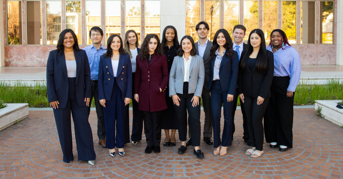 A group photo of twelve smiling graduates from the UCLA Executive MPH Class of 2026 in Health Policy and Management standing together outdoors on a brick courtyard in front of a campus building, dressed in professional business attire.