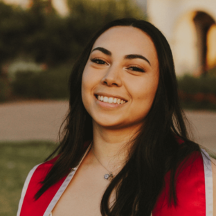 Headshot of Juliane Martinez, a student in the UCLA Executive MPH program Class of 2028, with long, straight brown hair, smiling outdoors in a red graduation stole