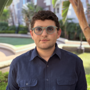 headshot of UCLA Executive MPH student Miller Finkelstein wearing a dark blue button front collared shirt with front pockets. He has short brown hair and he is wearing round rimmed sunglasses. He stands outside with trees behind him